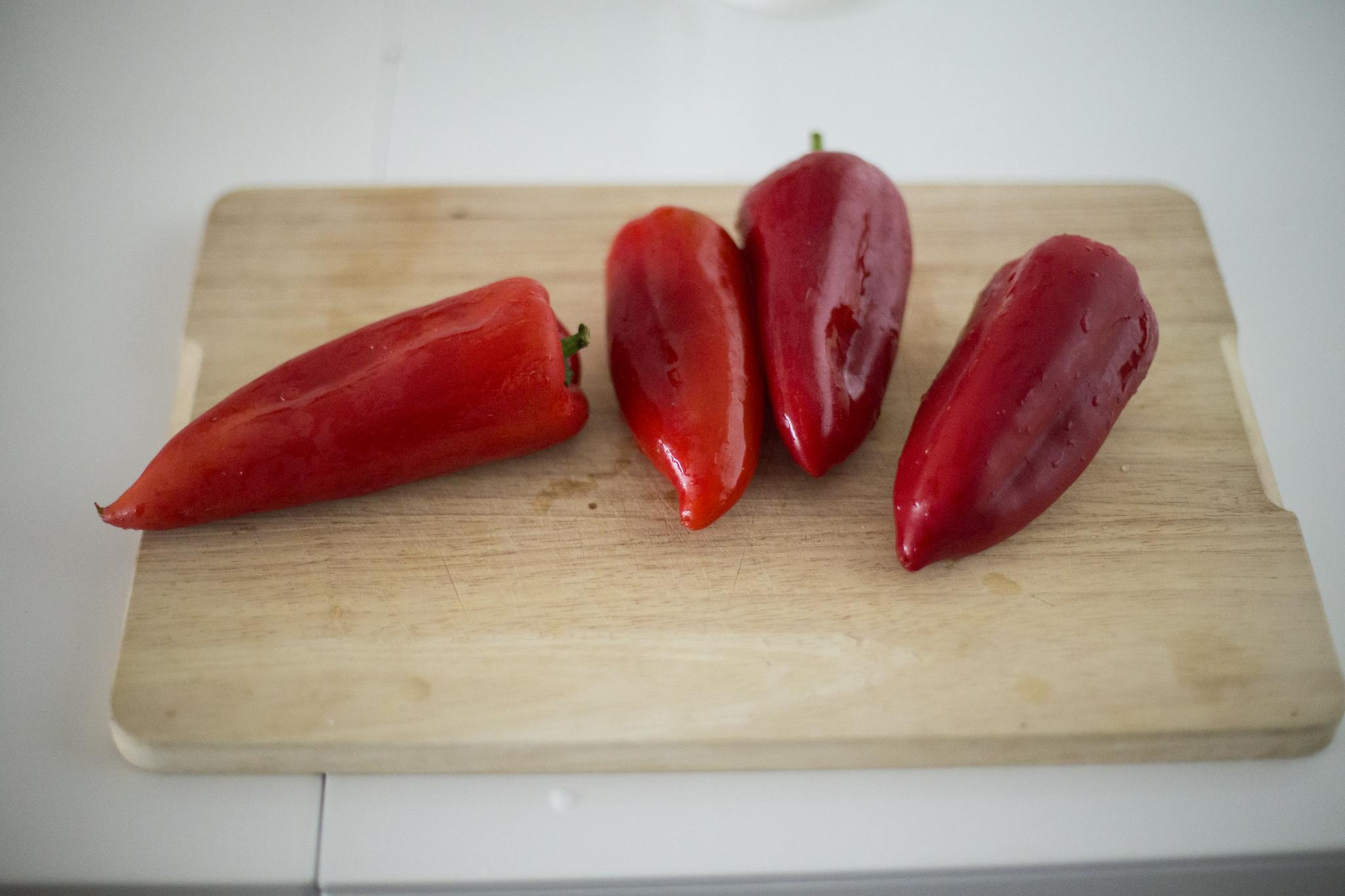 Four fresh red peppers displayed on a wooden chopping board in natural light.