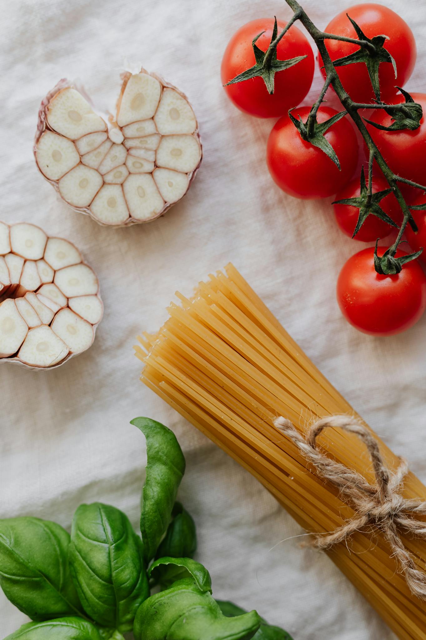 Top view of fresh tomatoes, garlic, basil, and uncooked pasta on a white background.